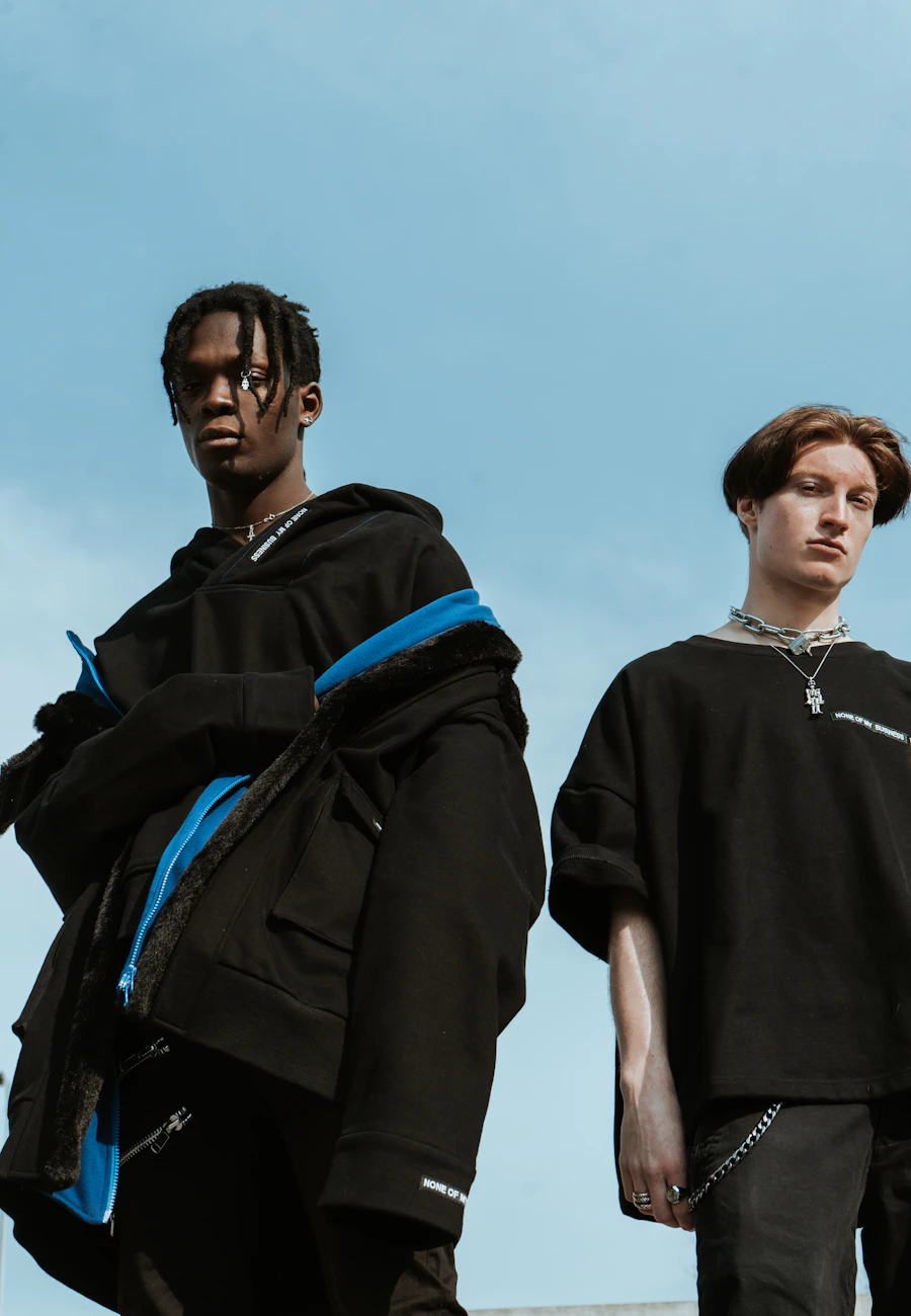 Two young men in black technical jackets and oversized tees against a blue sky